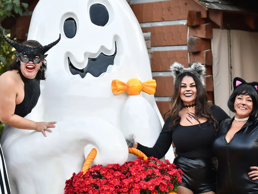 Three guests in black costumes with cat ear and horn head bands laugh beside a large white ghost statue.