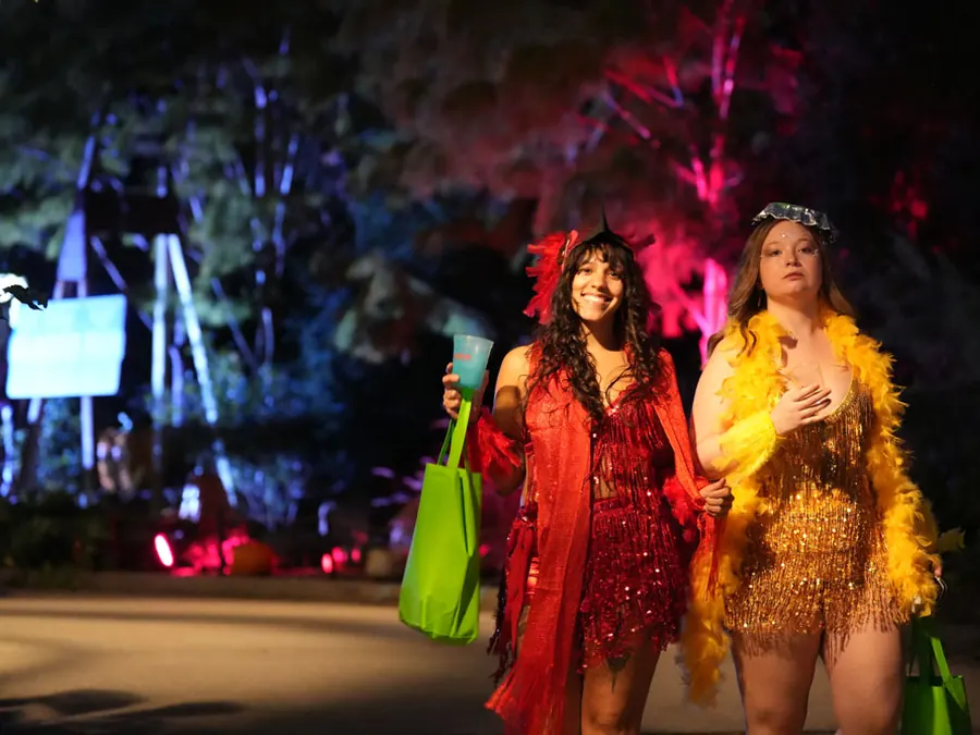 Two guests in red and gold sequined costumes with feather boas walk a zoo path lit by pink and blue lights at night.