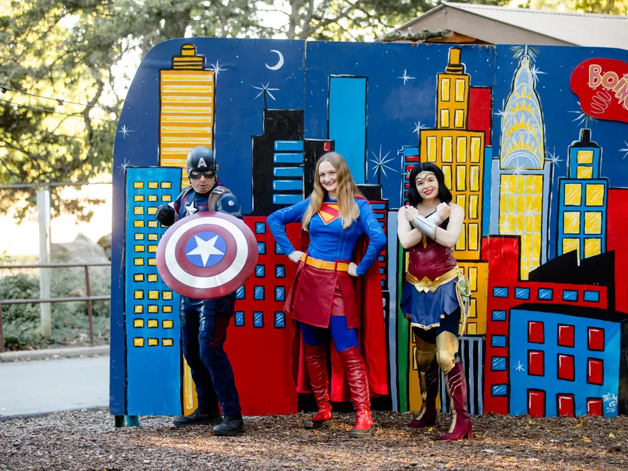 Guests dressed as Captain America, Supergirl, and Wonder Woman pose in front of a painted city skyline backdrop.