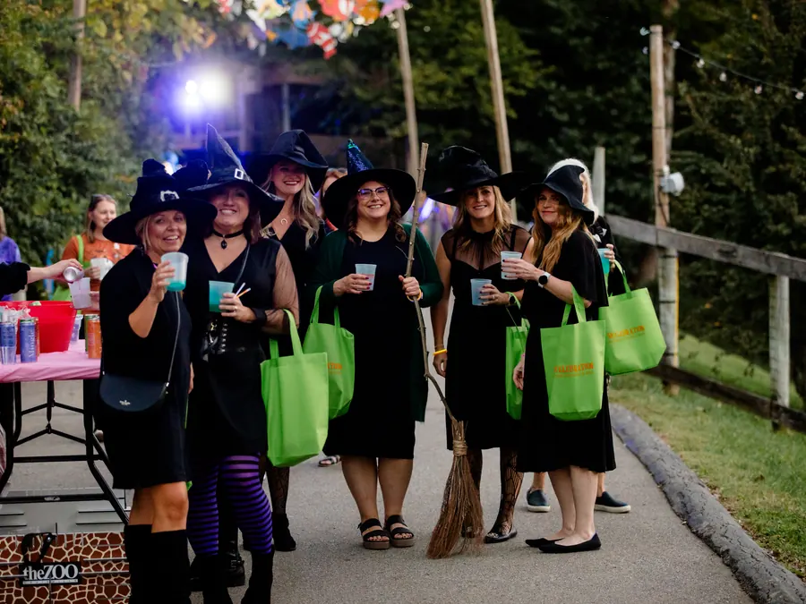 Six guests in black witch hats and dresses hold drinks and green Skelebration tote bags on a zoo path at dusk.